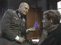 Oberleutnant Kluge (John Malcolm) questions Peter Porteous (Richard Heffer) about the appearance of paper "V"s., bookmarking the island's library books.